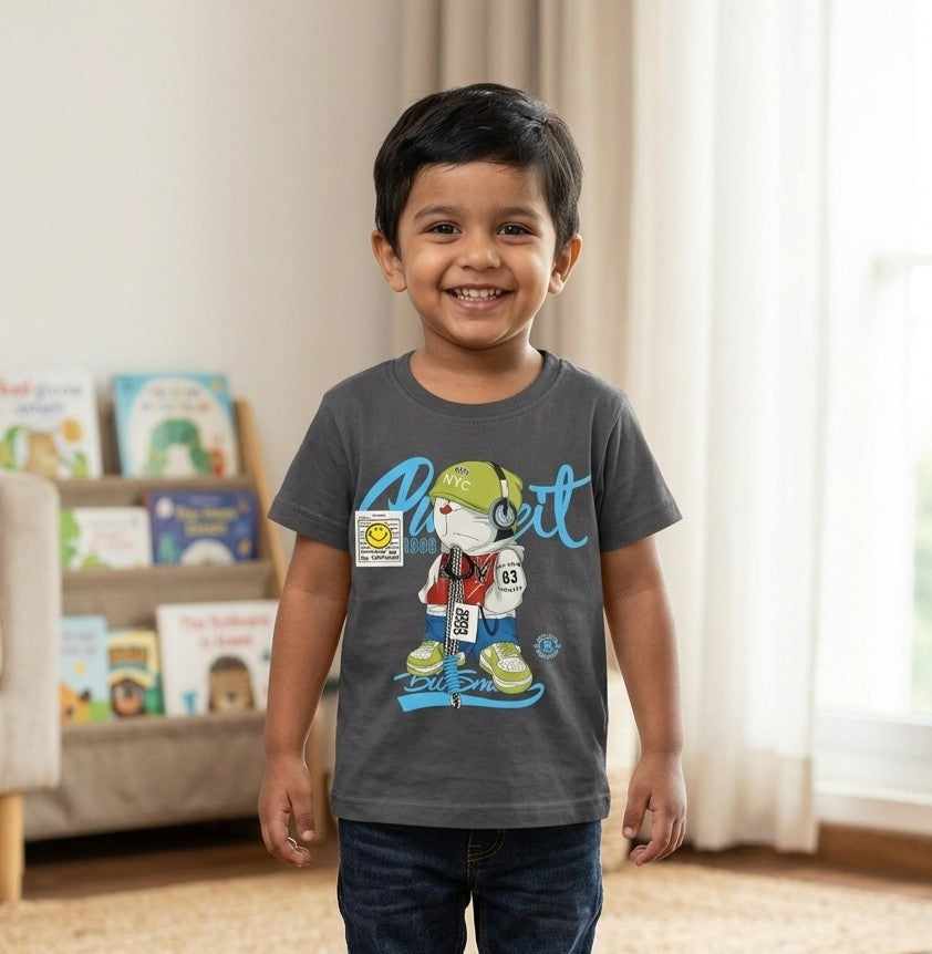 A kid wearing a FYST brand charcoal grey short-sleeved t-shirt featuring a large streetwear-inspired graphic of a character with headphones, blue script lettering, and a small yellow smiley face badge detail.