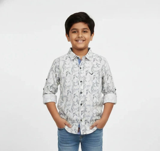 A young boy smiling with his hands in his pockets, modeling a Fyst brand light grey button-down shirt featuring an all-over delicate leaf and branch print. The shirt includes contrasting dark buttons, a small chest logo, and sleeves with roll-up tab details, set against a solid white background.