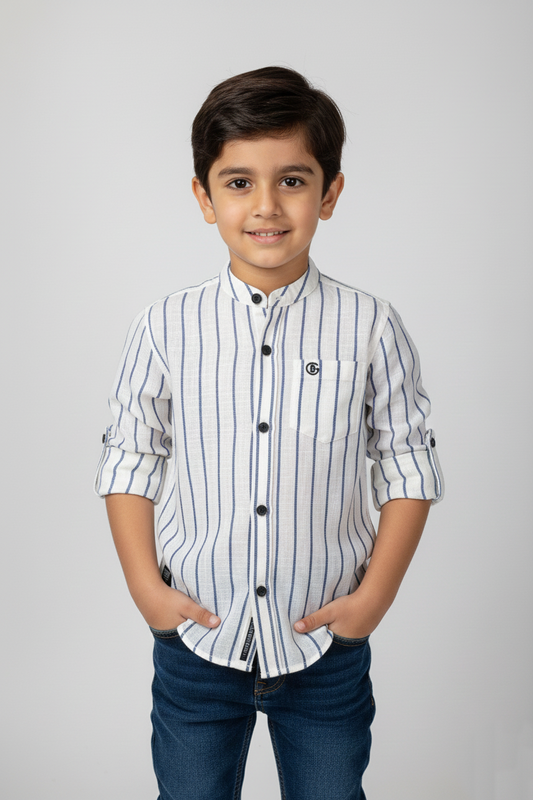 A front-facing photograph of a smiling young boy wearing a Fyst brand off-white, long-sleeve linen-texture button-down shirt with a mandarin collar. The shirt features thin vertical navy pinstripes, black buttons, a single chest pocket with a small 'G' emblem, and rolled-up sleeves. The boy has his hands in the pockets of dark blue jeans, posed against a solid light grey studio background. 