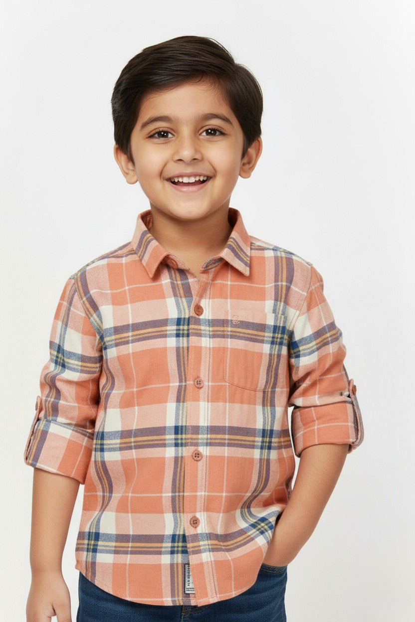 A cheerful young boy wearing a Fyst brand long-sleeve button-down shirt featuring a peach, cream, and navy blue plaid check pattern. The shirt has a classic collar, wood-style buttons, and sleeves rolled up with button tabs. He is smiling and posed against a clean white background, showcasing the shirt's vibrant pattern and soft cotton texture.
