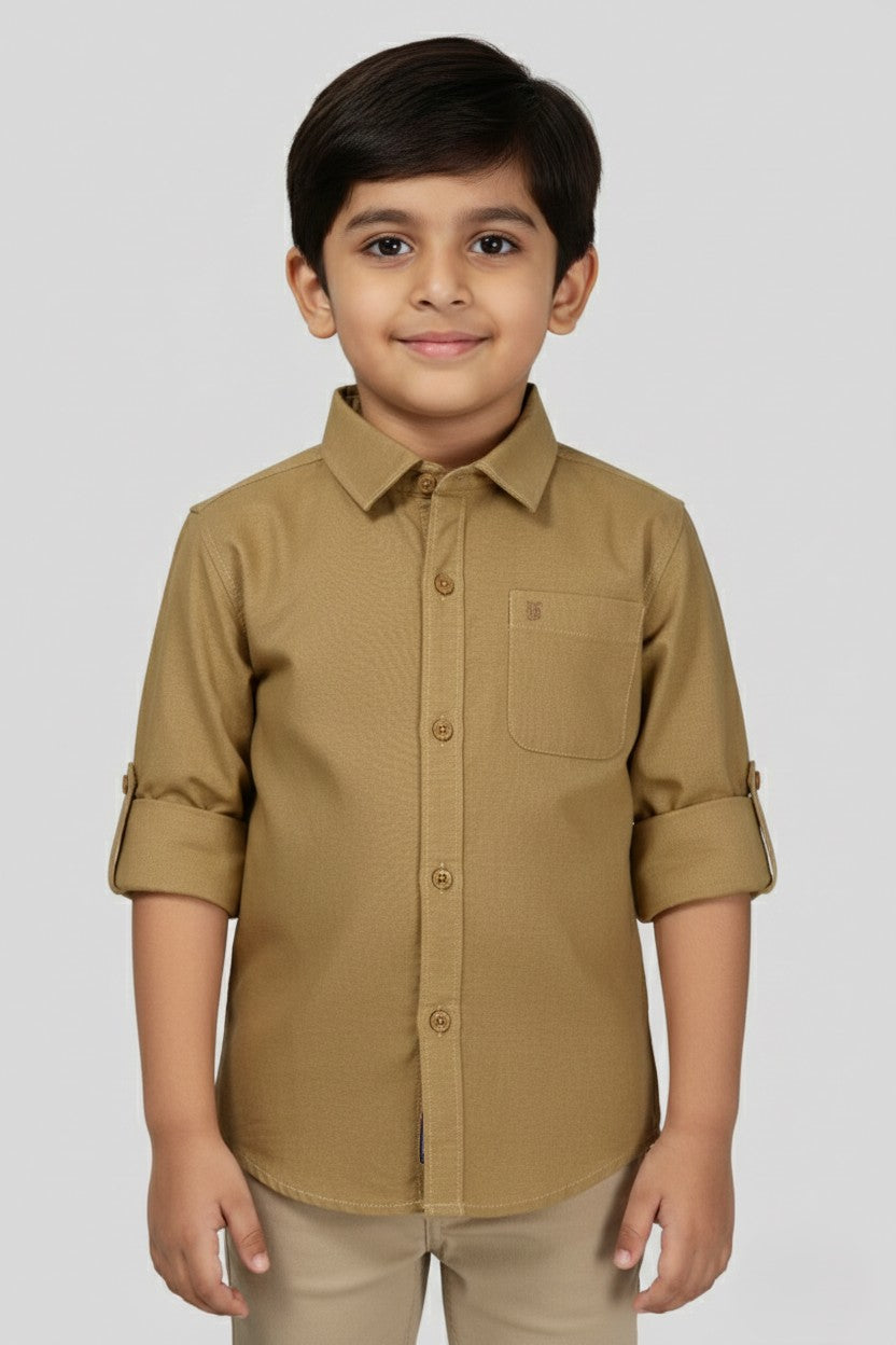 A young boy models a Fyst brand solid khaki beige long-sleeve cotton shirt. The shirt features a classic spread collar, wood-tone buttons, a single chest pocket, and sleeves neatly rolled up with button tabs. He is posed against a neutral grey background, highlighting the shirt's clean tailoring and versatile earth-tone color.