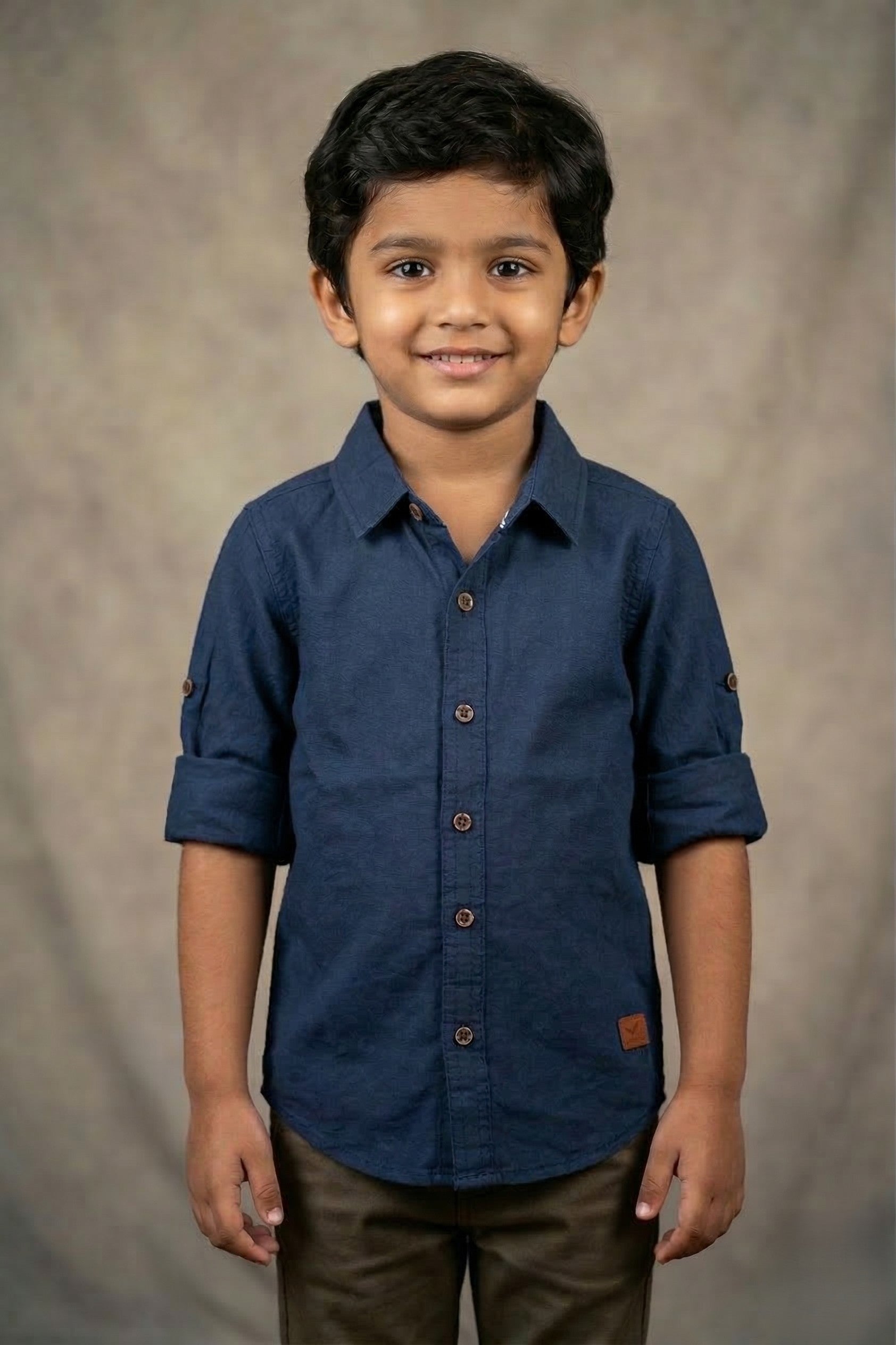 smiling young boy models a Fyst brand navy blue long-sleeve cotton shirt with a textured finish. The shirt features a classic spread collar, contrast brown buttons, and sleeves rolled up to the elbow with button tabs. He is wearing olive green trousers and posed against a mottled tan studio background, highlighting the shirt's rich color and smart-casual design.