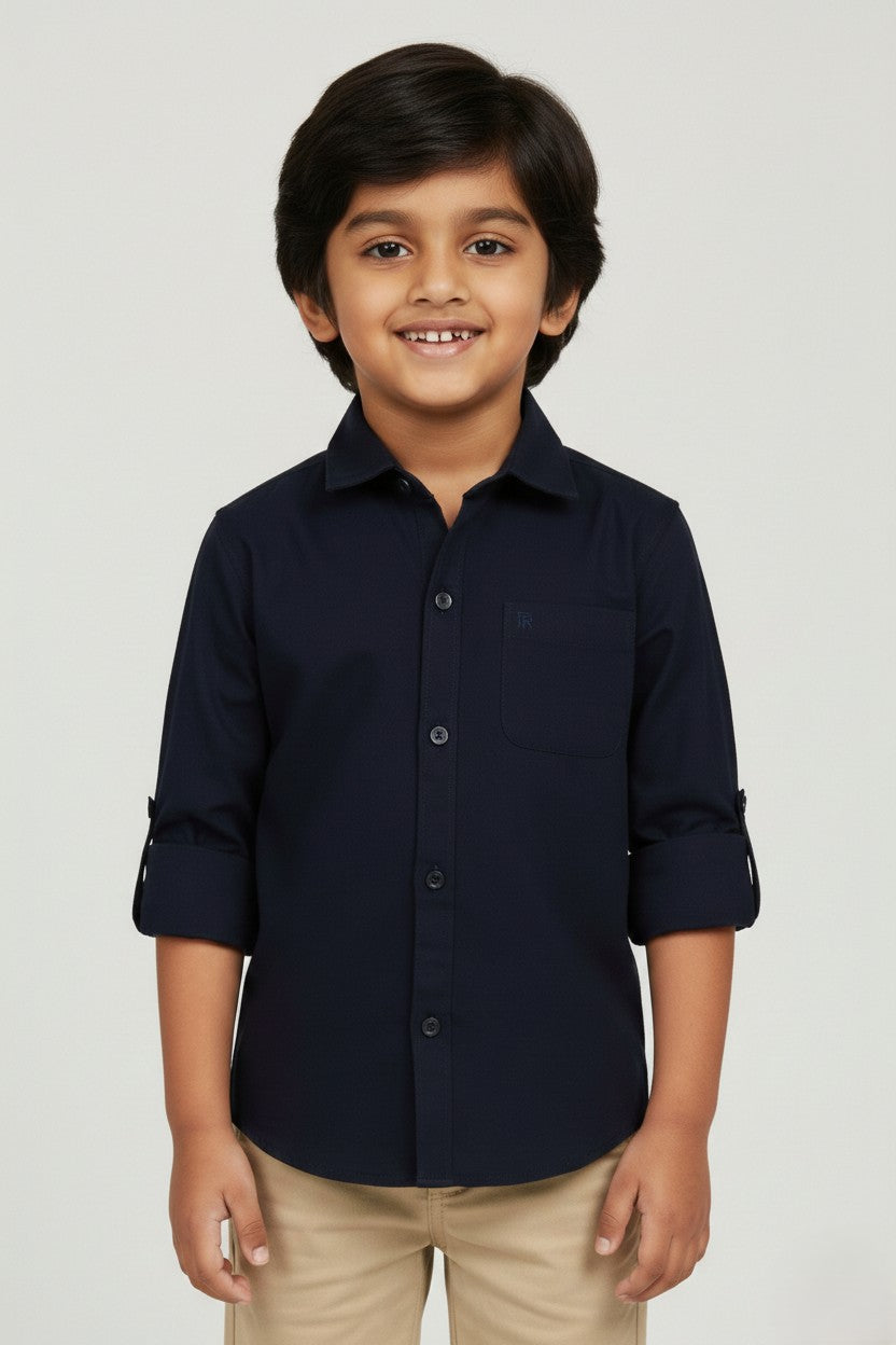 A smiling young boy wearing a Fyst brand solid navy blue long-sleeve cotton shirt. The shirt features a classic spread collar, matching navy buttons, a single chest pocket, and sleeves rolled up with button tabs. He is paired with khaki trousers and posed against a light grey background, showcasing the shirt's sharp tailoring and deep, professional blue color.