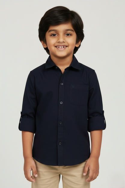 A smiling young boy wearing a Fyst brand solid navy blue long-sleeve cotton shirt. The shirt features a classic spread collar, matching navy buttons, a single chest pocket, and sleeves rolled up with button tabs. He is paired with khaki trousers and posed against a light grey background, showcasing the shirt's sharp tailoring and deep, professional blue color.