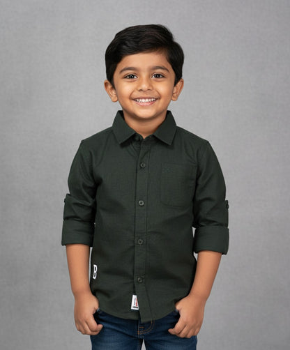 A smiling young boy models a Fyst brand solid olive green long-sleeve cotton shirt. The shirt features a classic spread collar, color-matched dark buttons, a single chest pocket, and sleeves rolled up with button tabs. He is posed against a textured grey background, showcasing the shirt's rich earth-tone color and durable fabric.