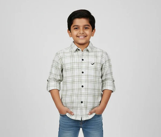 A young boy smiling with his hands in his pockets, modeling a Fyst brand off-white button-down shirt featuring a green and grey windowpane check pattern. The shirt has dark contrasting buttons, a small black logo on the chest, and long sleeves with roll-up tab details, shown against a neutral studio background.