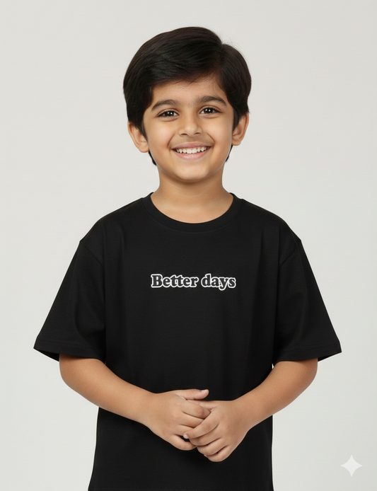 A young boy modeling a black oversized t-shirt. The front features a small neutral-toned graphic on the chest, while the back displays a large, intricate streetwear-style typography design. He is posing against a plain grey background.