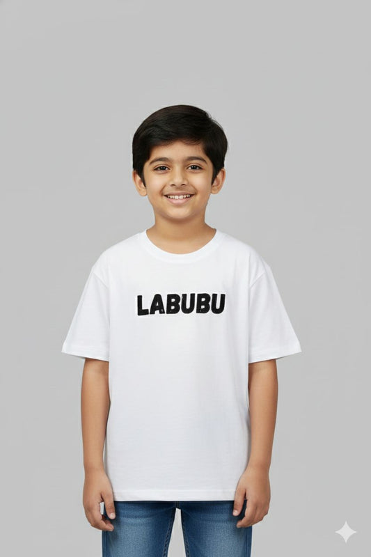 A young boy modeling white oversized t-shirt. The front features a small neutral-toned graphic on the chest, while the back displays a large, intricate streetwear-style typography design. He is posing against a plain grey background.