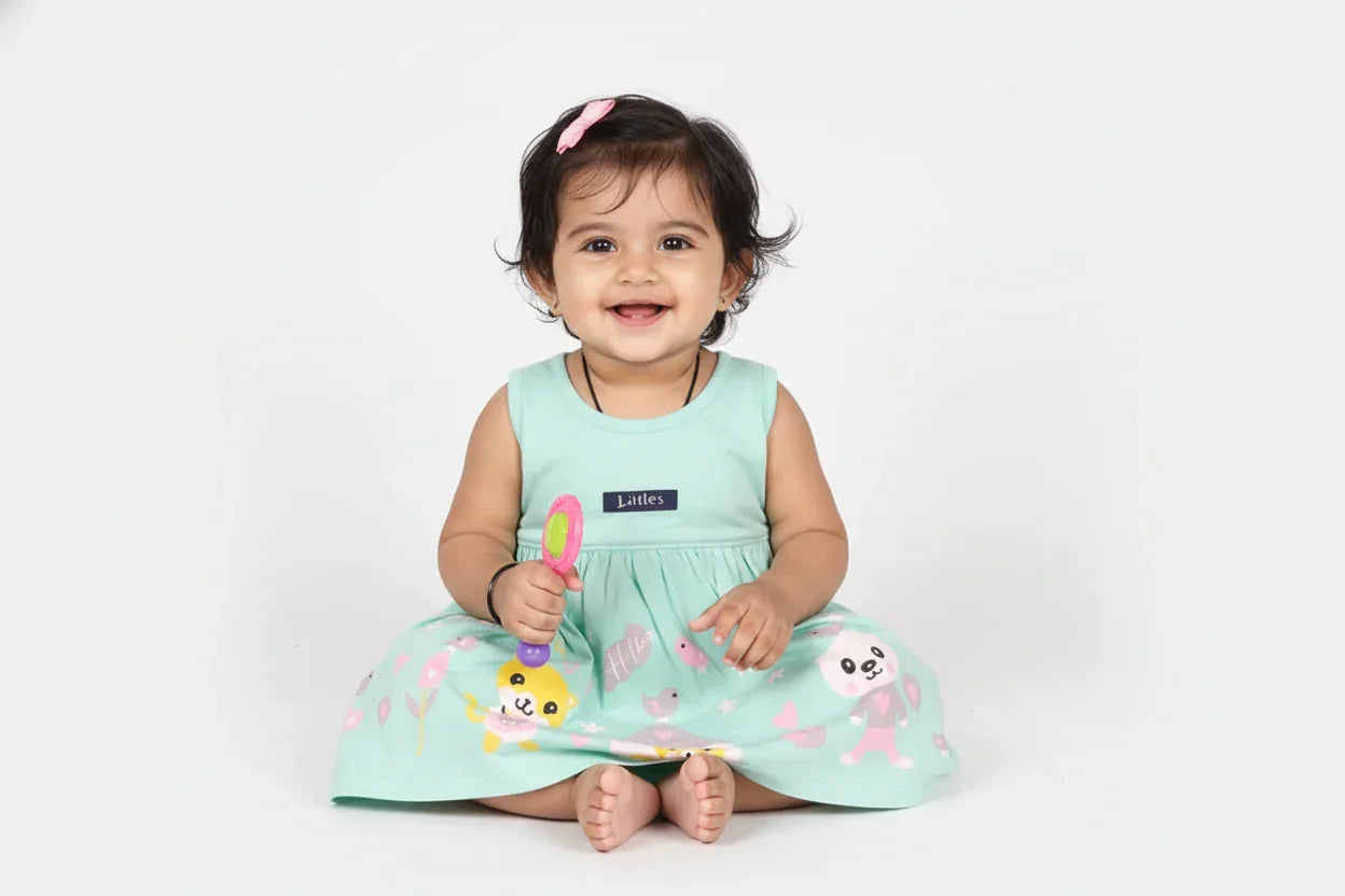 Smiling baby girl in sea green cotton knit frock with animal graphics, sitting and holding a toy