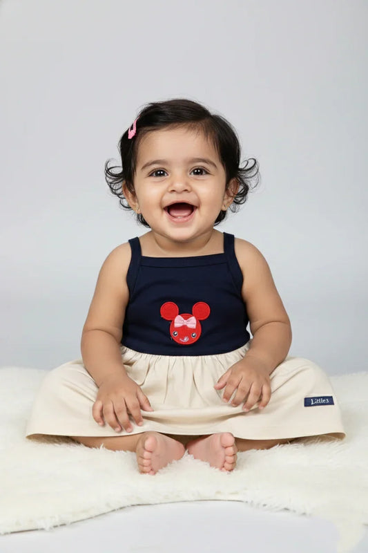 Smiling baby in navy and cream cotton knit strappy frock with cartoon patch, sitting on white fur