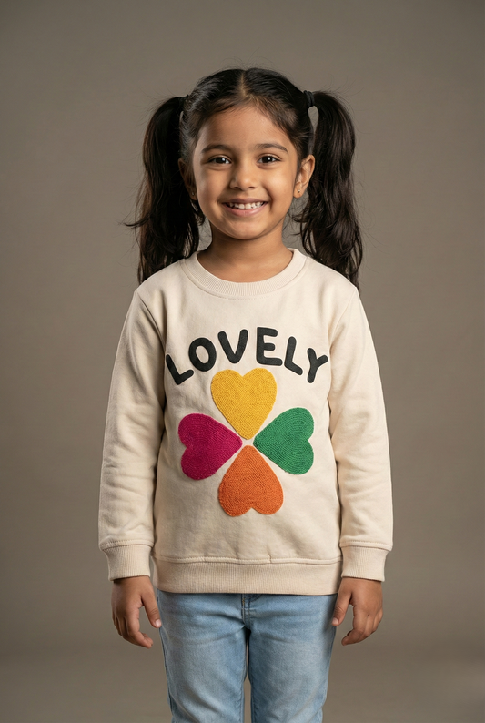"A professional product showcase of the Fyst girls' cream-colored graphic sweatshirt. The primary image features a young girl with pigtails modeling the soft fleece top, which displays the word 'LOVELY' in bold black capital letters above a large four-leaf clover graphic composed of yellow, green, red, and orange hearts. Detailed thumbnails provide a 360-degree view, highlighting the clean back profile, the high-quality ribbed texture of the crew neckline and cuffs, and a close-up of the vibrant chest graph