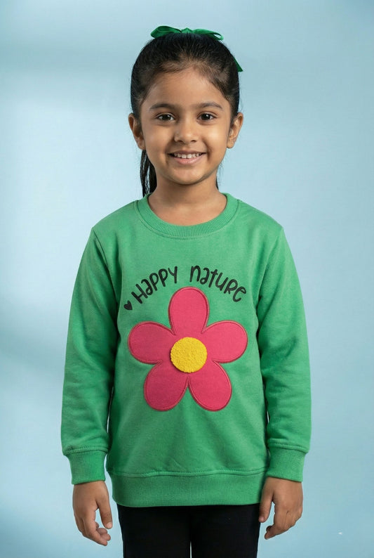 A bright green long-sleeved sweatshirt for children. It features the text 'Happy nature' printed in black above a large, fabric appliqué of a flower with five hot pink petals and a textured yellow center. The sweatshirt includes a ribbed crew neck, ribbed cuffs, and a ribbed hem