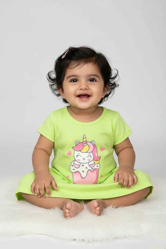 Baby girl in green cotton frock with unicorn graphic, sitting and smiling on white fur rug