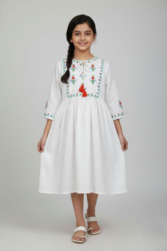 A white, long-sleeved dress for a young girl hanging on a wooden hanger. It features a square bodice section with red and blue floral embroidery and green vine details. The dress has a round neckline with a tie closure ending in orange tassels, elasticated cuffs with matching embroidery, and a gathered, flowy skirt
