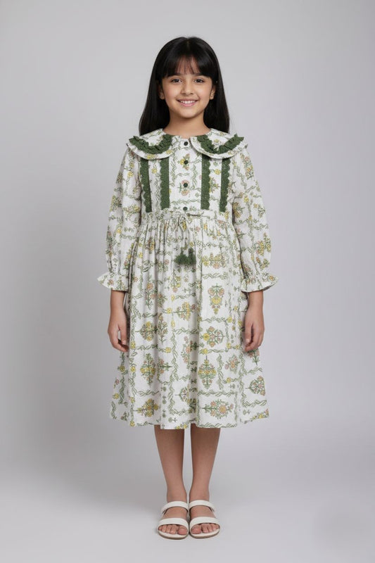 A white long-sleeved dress for a young girl featuring a delicate yellow and pink floral print. The dress has a Peter Pan collar and bodice detailed with dark green lace trim, a button-down front, and a drawstring waist with green tassels. It is hanging on a wooden hanger