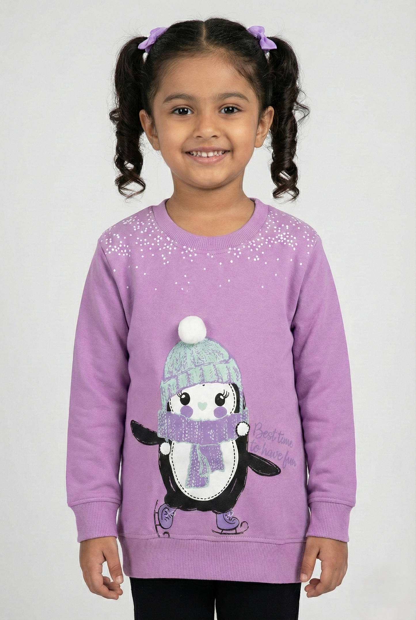 Purple long-sleeve crewneck sweatshirt for girls. The center features a large graphic of a cute cartoon penguin ice skating, wearing a mint green knit hat with a white pom-pom and a purple scarf. White speckled snow details appear near the neckline, and the text 'Best time to have fun' is printed on the side