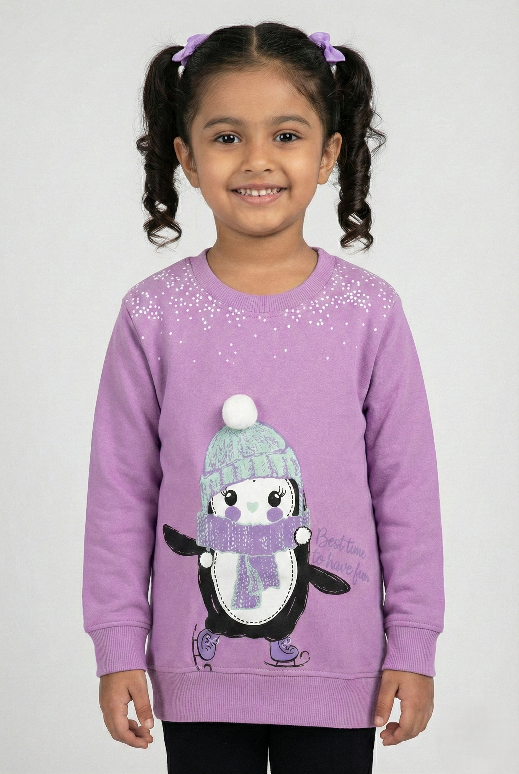 Purple long-sleeve crewneck sweatshirt for girls. The center features a large graphic of a cute cartoon penguin ice skating, wearing a mint green knit hat with a white pom-pom and a purple scarf. White speckled snow details appear near the neckline, and the text 'Best time to have fun' is printed on the side